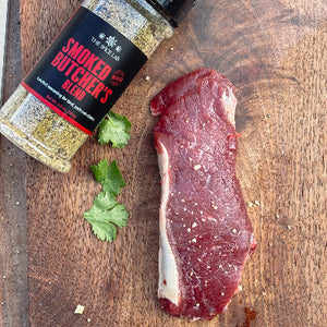 Spice bottle labeled 'Smoked Butcher's Blend' next to a piece of raw elk meat on a wooden surface.