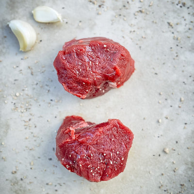 Two pieces of raw elk filet on a textured surface with garlic cloves.