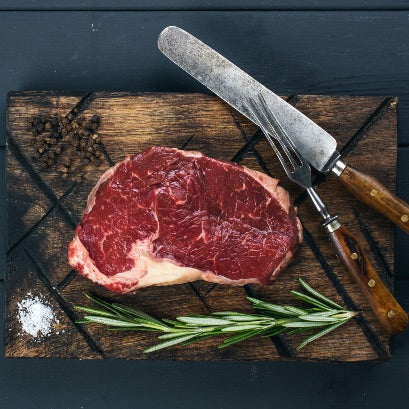 Bison steak on a wooden cutting board with a knife and fork, surrounded by rosemary and salt.